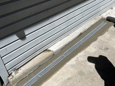 A concrete path with a metal drainage grate runs alongside a vinyl-sided building. A shadow of a person is visible on the right.