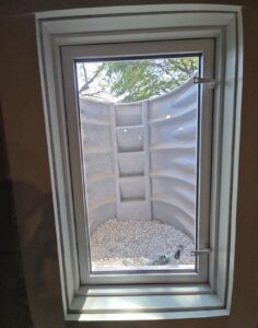 A window at basement level shows an exterior window well with gravel at the bottom and a view of tree branches above.