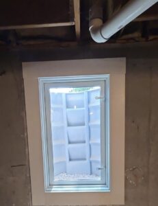 A small basement window with a white frame is set in a concrete wall, looking out to a window well with a corrugated metal barrier and some gravel at the bottom. A pipe runs along the ceiling.