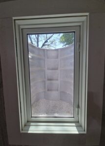 A basement window with a white frame looks out onto a window well lined with gravel and surrounded by corrugated gray panels; trees are visible at the top.