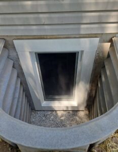 A basement window with a white frame is set in a concrete foundation, surrounded by a metal window well and gravel at the base.