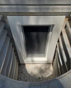 A basement window with a white frame is set in a concrete window well, surrounded by corrugated metal and gravel at the bottom.