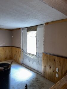 A room with wood paneling, beige walls, an unfinished window area with exposed drywall and plaster, and a trash bin in the foreground.
