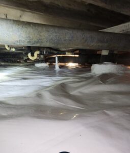 View of a crawl space with exposed pipes and beams above a white plastic vapor barrier covering the ground. Light is visible in the background.