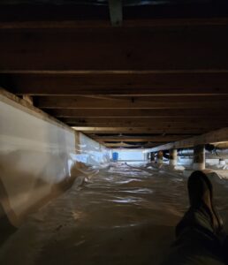 Person lying down inside a crawl space with a plastic vapor barrier covering the ground and lower walls, wooden beams above, and light visible at the far end.