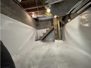 A basement with exposed ceiling beams and pipes, lit by bulbs, has walls and floor covered in white plastic sheeting, with wooden stairs leading up.