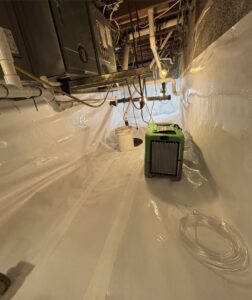 A basement or crawl space with white plastic lining on the floor and walls, a green dehumidifier, a white bucket, exposed pipes, and ducts visible overhead.