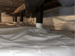 Plastic sheeting covers the ground and walls in a crawl space, with black pipes running overhead and seams sealed with white tape.