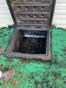 An open plastic access hatch set into the ground next to a building, showing a dark chamber below. Green grass seed and soil cover the surrounding area.