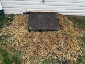 A plastic basement hatch is surrounded by straw on the ground next to the exterior wall of a house.