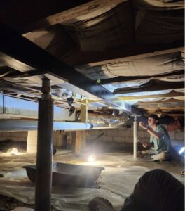 A person works under a house in a crawl space with exposed insulation, pipes, and construction lights illuminating the area.