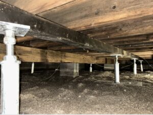 A crawl space under a house showing adjustable steel support posts, wooden beams, and a dirt ground.