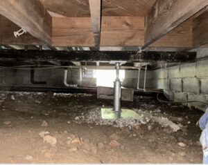 View of a crawl space with exposed soil, cinder block walls, wooden floor joists, pipes, and a metal support jack installed under the floor structure.