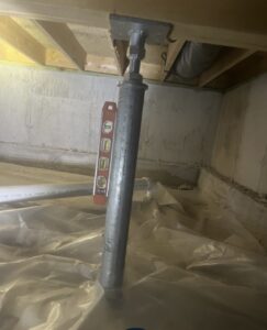 A steel adjustable support column is installed under a wooden floor with a level tool leaning against it in a basement or crawl space.