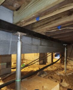 View of a crawl space showing metal support beams, adjustable steel columns, wooden floor joists, and utility lines running across a dirt floor.