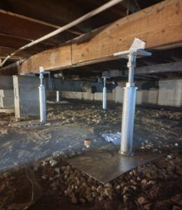 Steel support jacks installed beneath a wooden floor structure in a crawl space with a dirt and gravel ground.