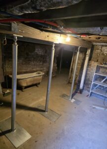 A dimly lit basement with metal support beams under wooden ceiling joists, exposed pipes, an old bathtub, and a shelf against brick and stone walls.