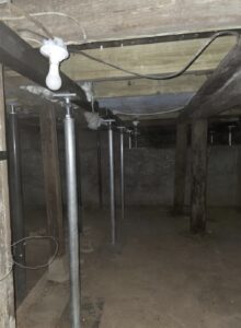A dimly lit basement or crawl space with exposed wooden beams, support columns, metal jacks, dirt floor, and a single hanging light bulb.