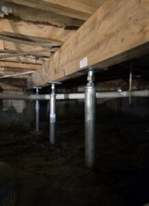 Metal support columns reinforce a wooden floor joist structure in a dimly lit crawl space with visible pipes and dirt ground.