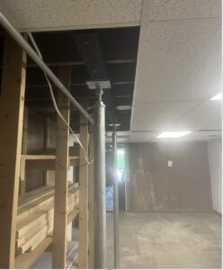A partially finished basement with exposed support beams and posts, wooden shelving on the left, and ceiling tiles, some of which are missing or misaligned.