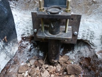 Close-up of metal foundation support post installed in concrete with bolts, surrounded by dirt and rocks.