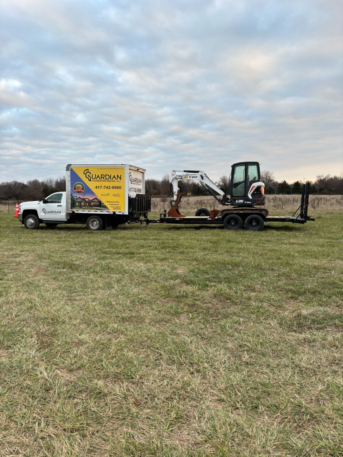 A white truck with a yellow logo and contact number is parked on grass, towing a flatbed trailer carrying a small excavator under a cloudy sky.