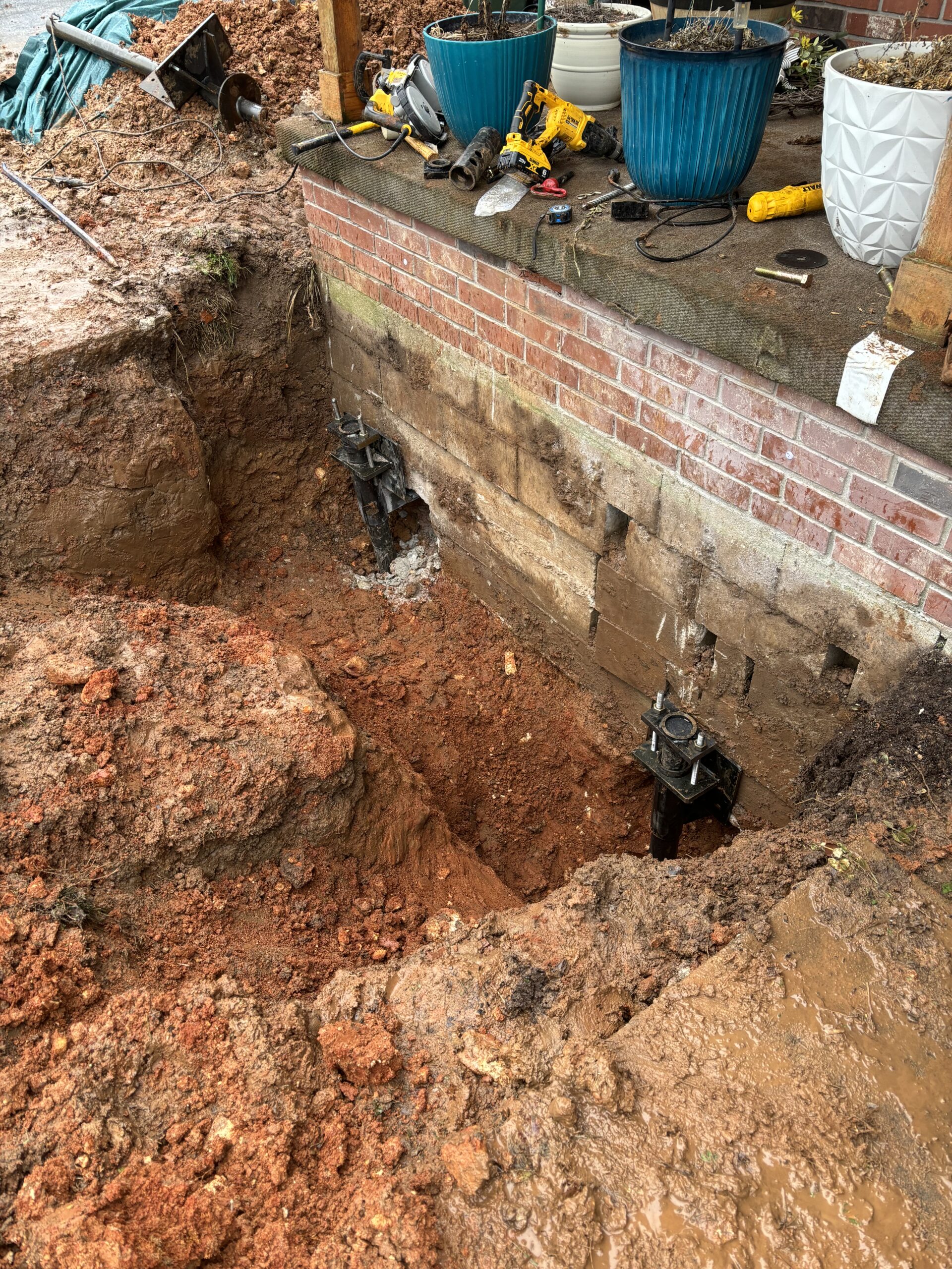 Excavation near a brick porch with two helical piers installed for foundation support; tools and plant pots rest on the porch above the exposed earth.