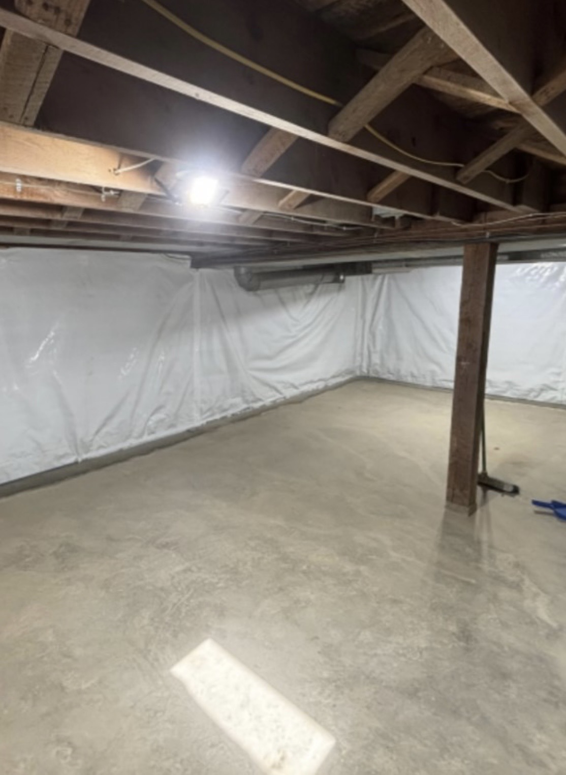 Unfinished basement with exposed wooden ceiling beams, concrete floor, and white plastic sheeting covering the walls. A support post stands near the center.