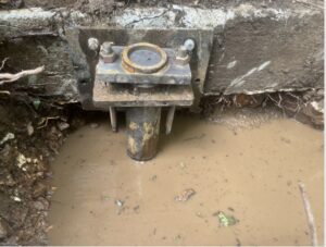 A metal foundation pier is installed against a concrete wall, partially submerged in muddy water and surrounded by wet soil.