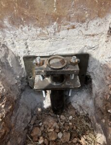 A steel foundation bracket is bolted to a concrete wall, anchored in an excavated dirt area with exposed soil and small rocks.