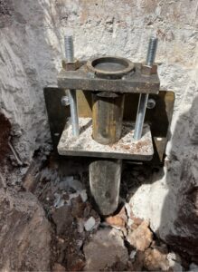 A metal pipe is secured in a concrete hole with a mounting bracket, bolts, and nuts. Broken pieces of concrete and debris surround the base.