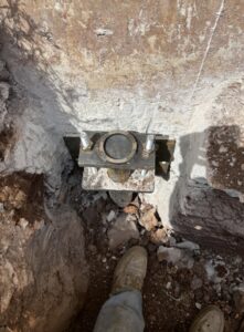 Metal foundation anchor plate with bolts embedded in concrete, surrounded by soil and rocks; part of a work boot is visible at the bottom of the image.