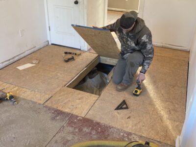 A person kneels on a wooden floor with a lifted panel, using a drill. A square tool and some screws are nearby. A door is in the background.