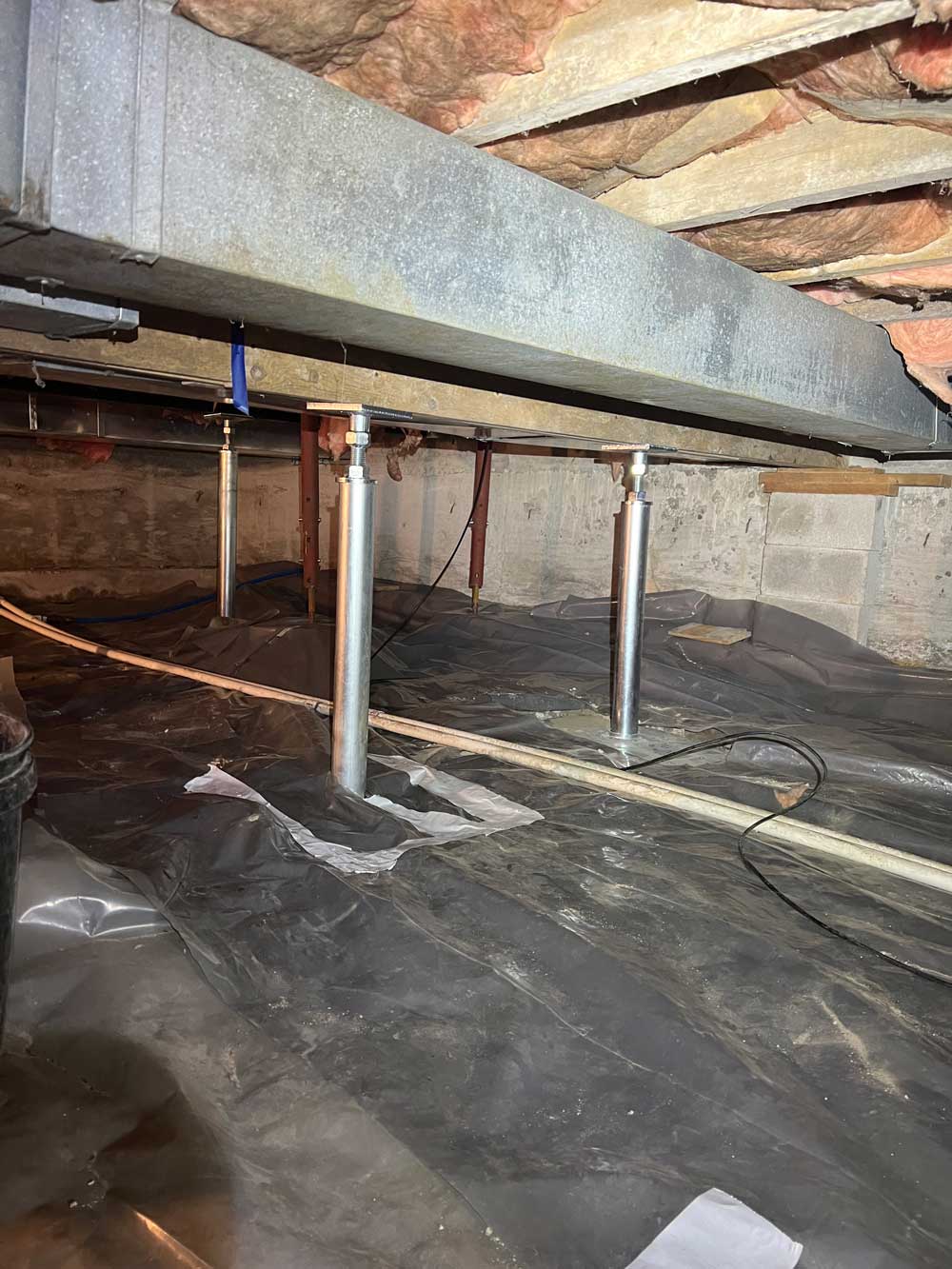 Crawl space with metal supports reinforcing beams, concrete walls, and insulation overhead. A plastic sheet covers the ground.