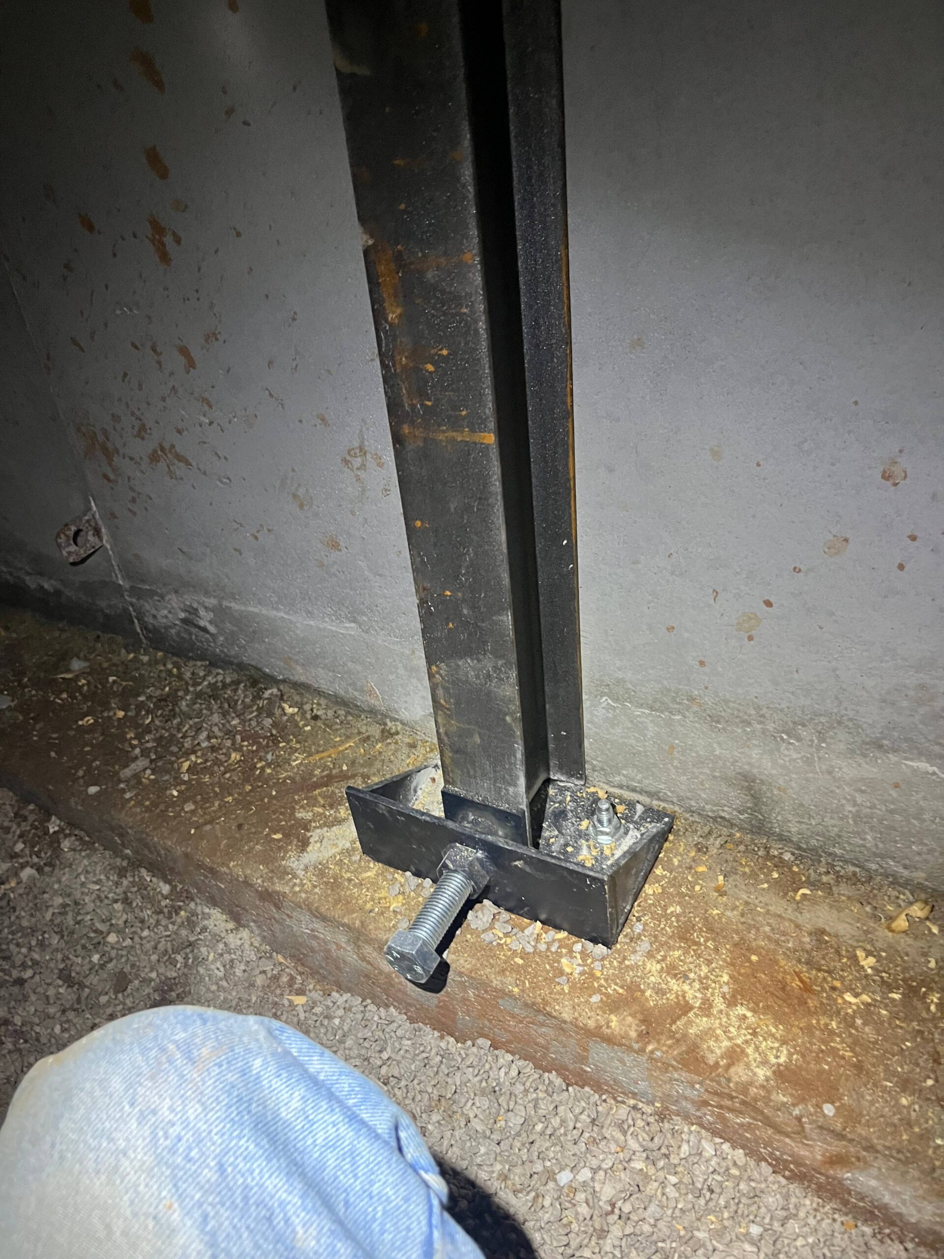 A metal post, possibly part of wall stabilizers, is anchored to a concrete surface with a square metal base plate and a large bolt; part of a person's knee is visible in the lower left corner.