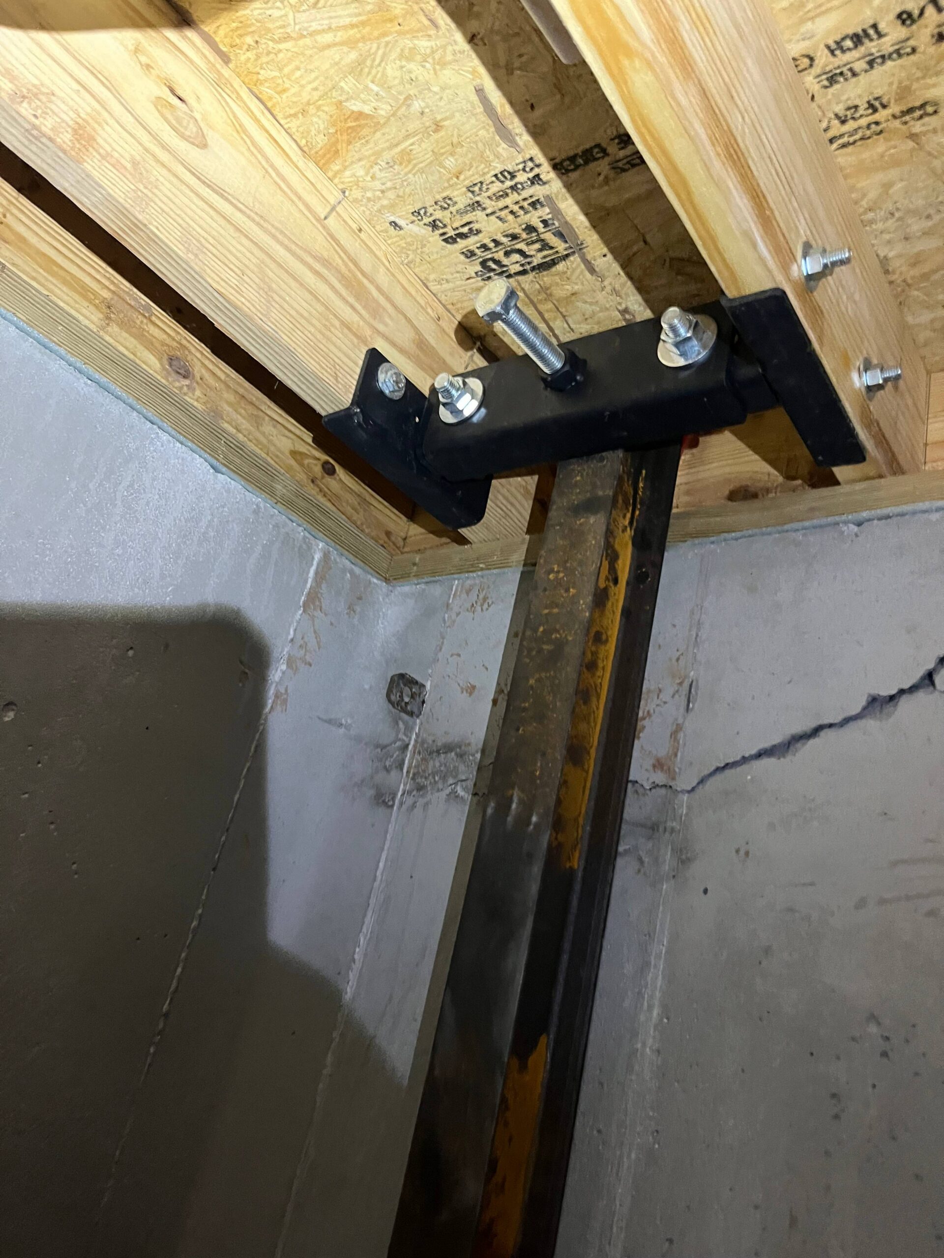A steel support post, serving as a wall stabilizer, is bolted to a wood beam and bracket, anchored to a concrete basement floor, showing structural reinforcement.