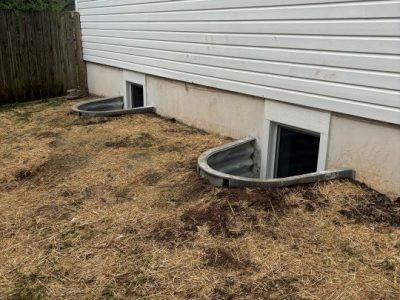 Two basement windows with curved metal window wells are set into the foundation of a building with white siding, surrounded by a patchy area of bare ground and straw.